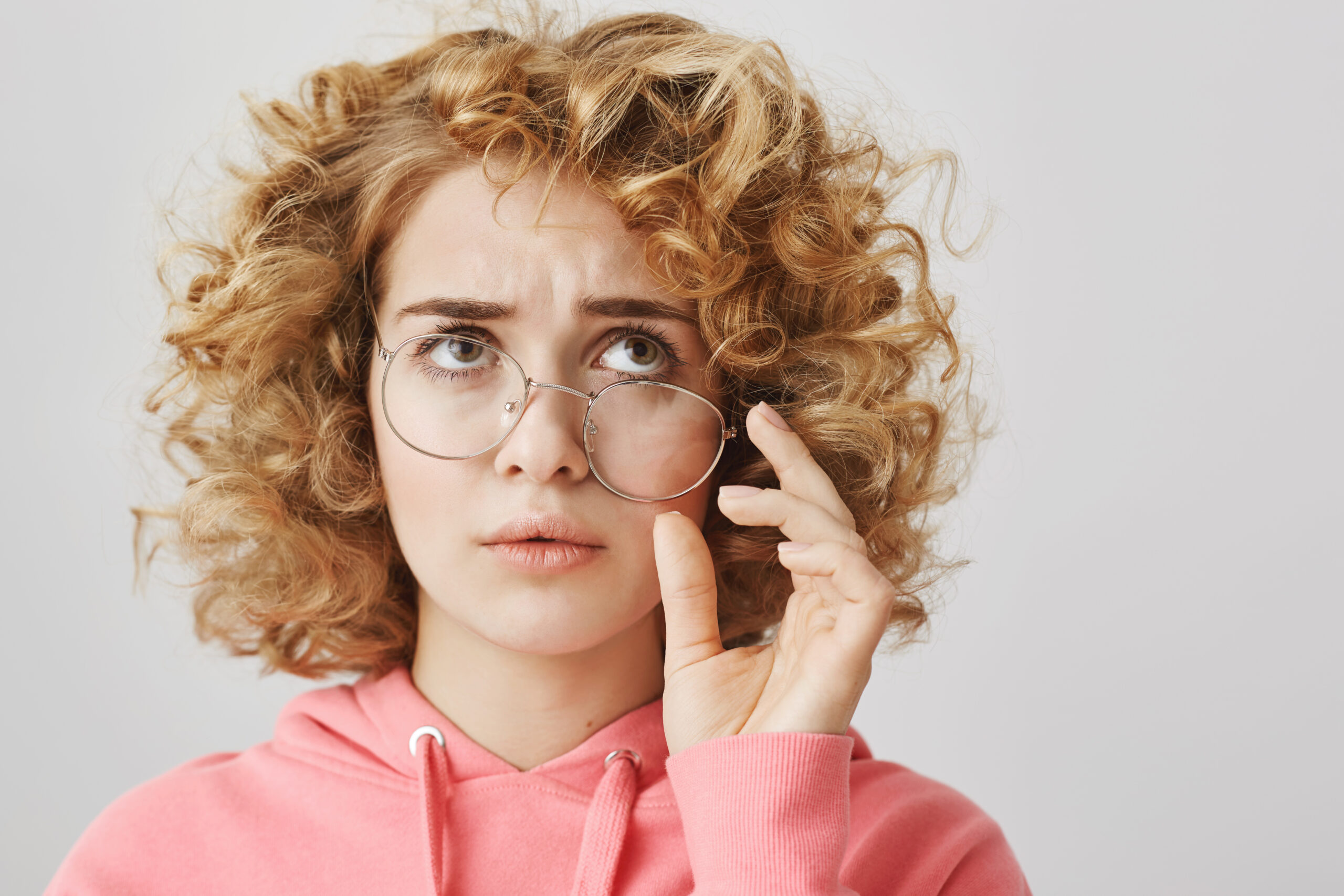 Confused and troubled woman tries to recall what happened. Portrait of worried and questioned curly-haired woman wearing crooked glasses, frowning and looking up, having no clue, posing over gray wall.